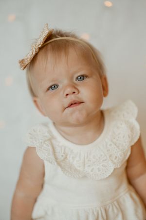 Baby in white lace dress with crown accessory against light backdrop with bokeh lights.