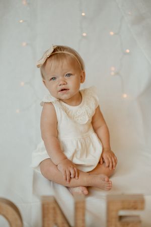 A baby in a white dress sits on the floor against a backdrop with soft twinkle lights.
