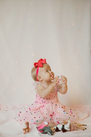 A baby in a pink dress sits among scattered confetti and small toys during a festive photo session.