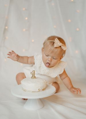Baby in white dress reaching for birthday cake with number 1 candle on white cake stand against twinkle light backdrop.