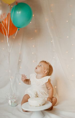 Baby in white dress sits by birthday cake with colorful balloons against sparkly backdrop during first birthday photo shoot.