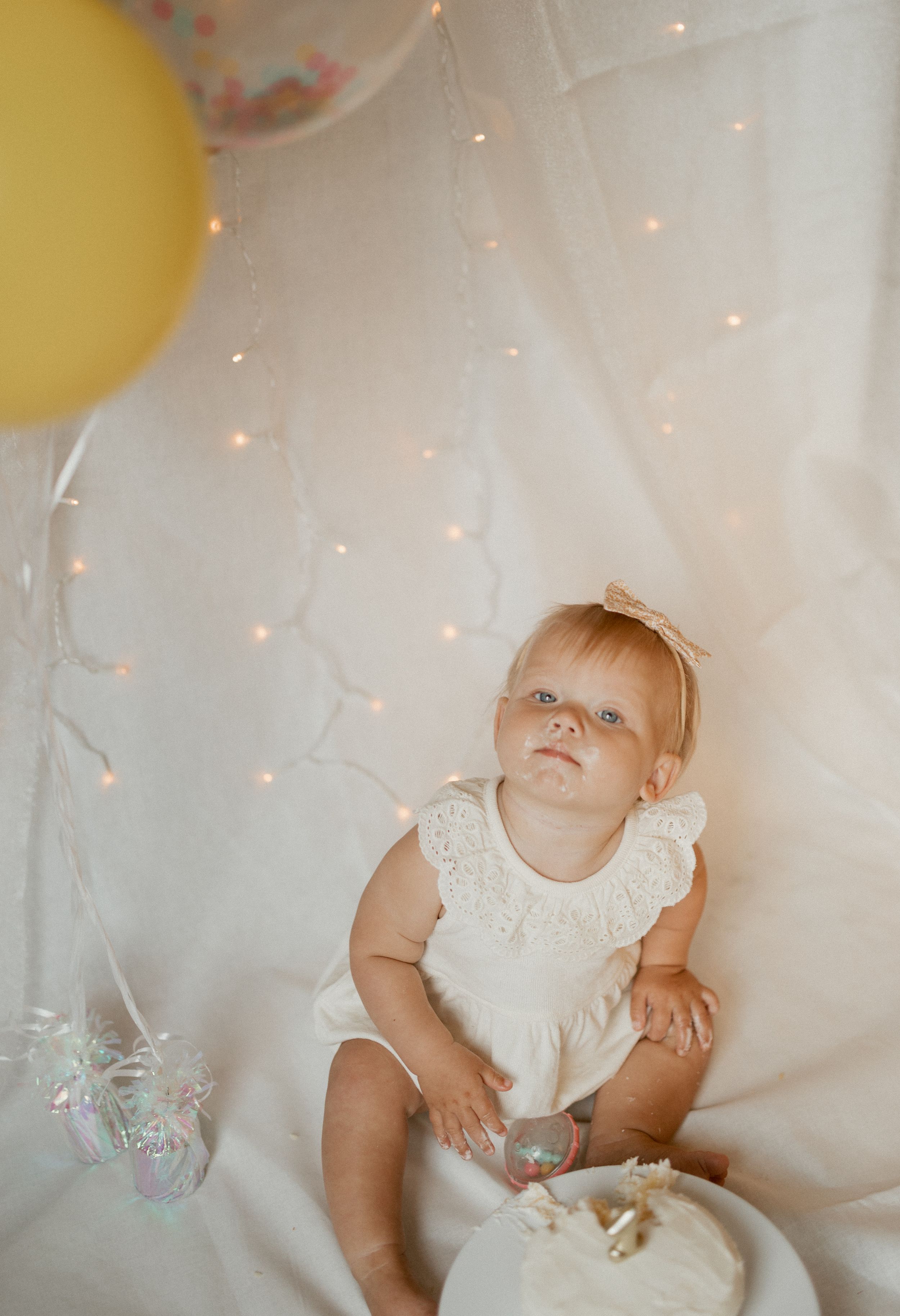 Baby wearing white lace dress sits on white blanket with balloon and twinkle lights in dreamy setting.