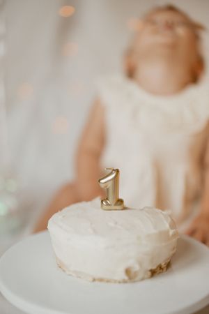 A simple white frosted cake with a gold number one candle on top sits on a white plate against a blurred background.