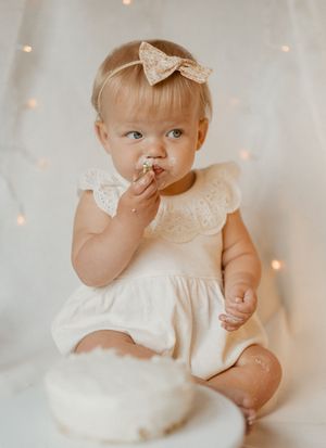 Baby wearing white lace dress and bow sits in front of twinkling lights while tasting something sweet.
