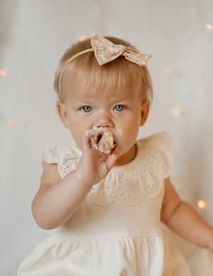 A small child in a white lace dress with a bow headpiece sits against a bokeh light background.
