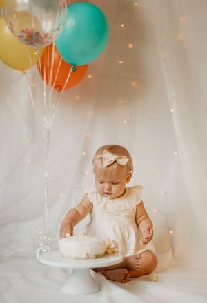 A baby in white dress sits beside a white cake during smash cake photoshoot with teal and yellow balloons.
