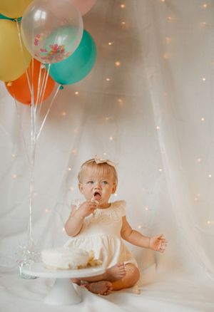 Baby in white surrounded by pastel balloons and twinkling lights for first birthday cake smash photo session.