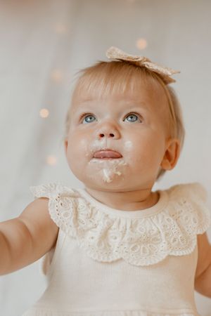 A young baby wearing a delicate white lace collar dress sits with sweet expressions in a soft light setting.
