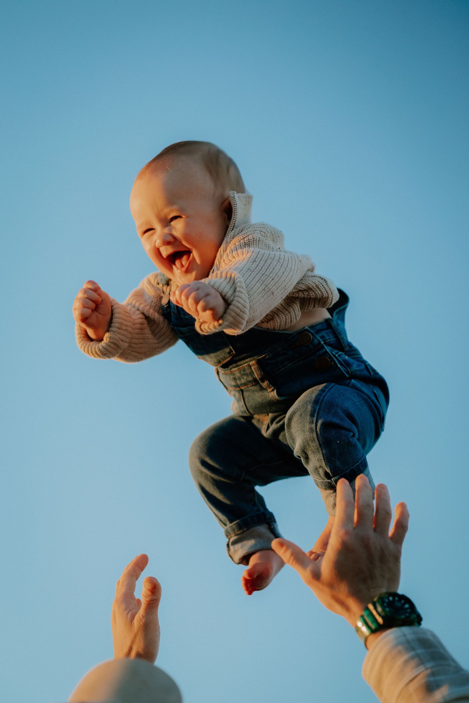 A baby wearing a sweater and jeans being tossed in the air against a blue sky, expressing pure joy and laughter.