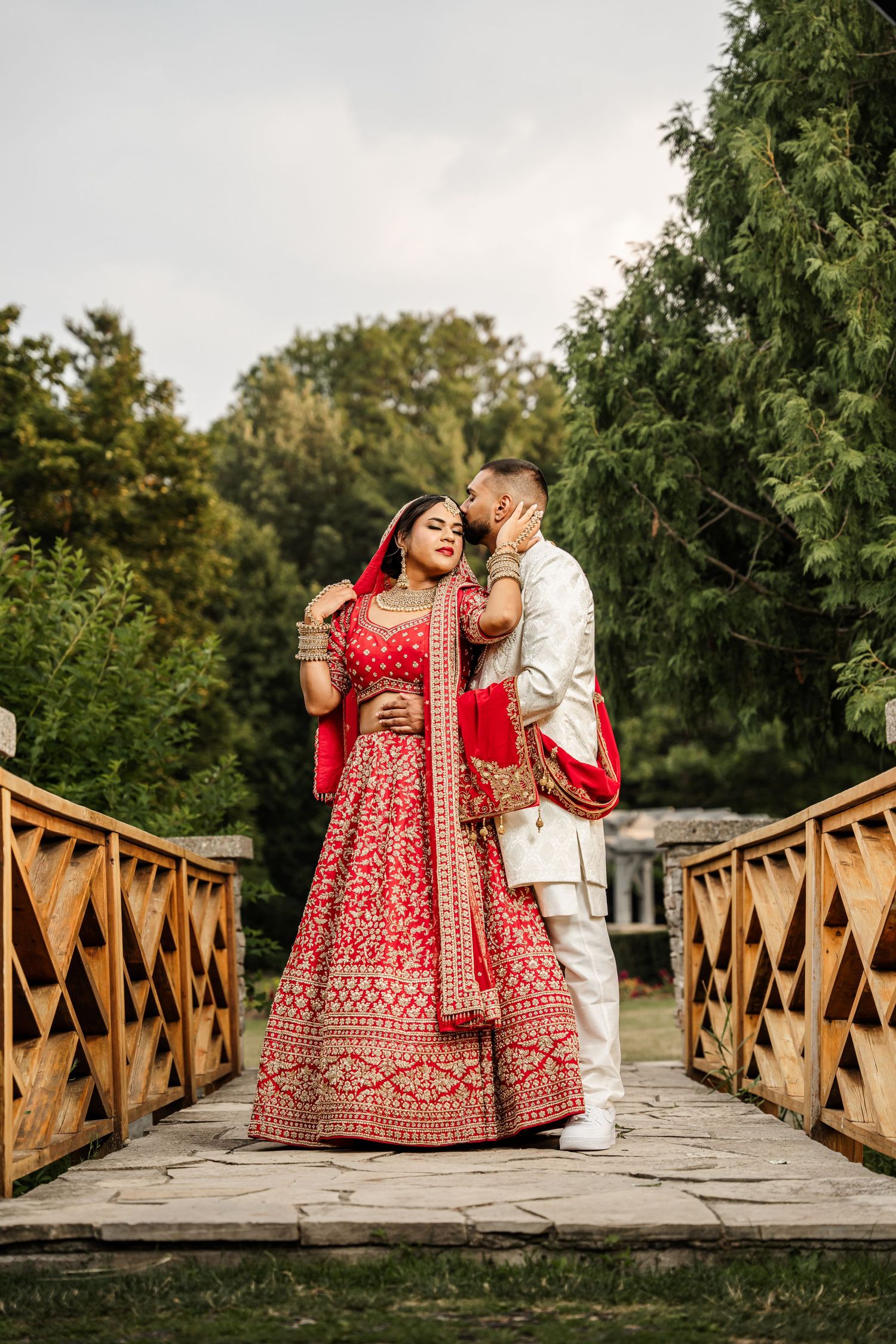 Traditional wedding couple in red lehenga and white sherwani pose on wooden bridge.