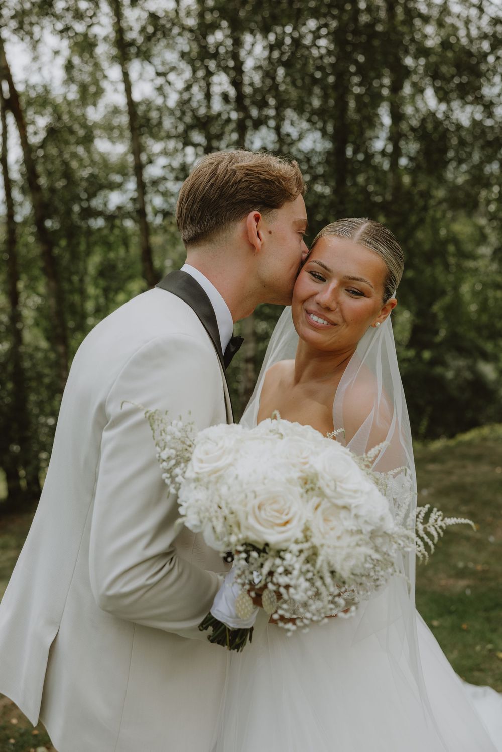 Bride holding white rose bouquet shares tender moment with groom in outdoor wedding setting.
