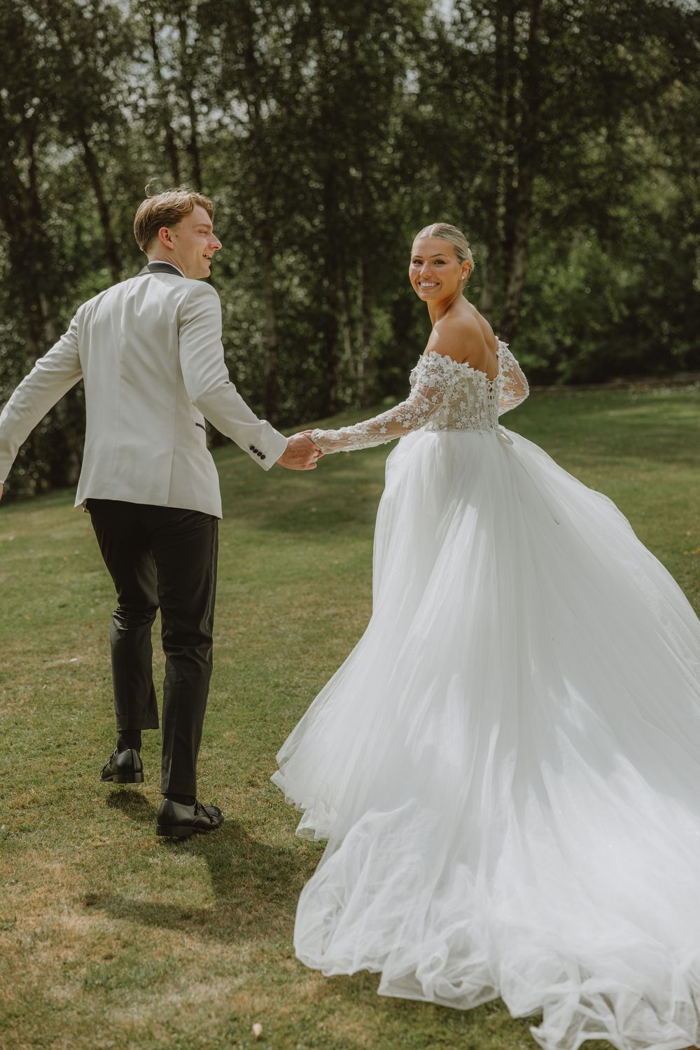 Couple in wedding attire dance on green lawn, bride wearing flowing lace ballgown.