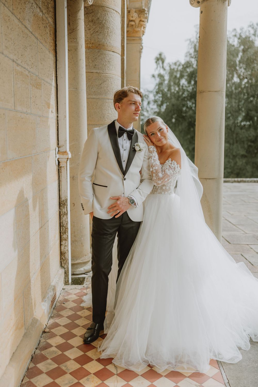 A wedding couple poses under elegant stone columns on a checkered floor.