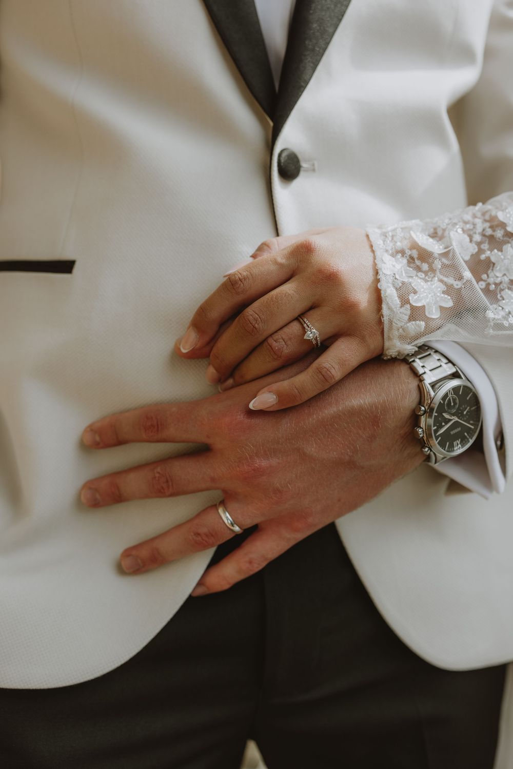 Close-up of newlyweds' hands showing wedding rings and a wristwatch against a white suit jacket.