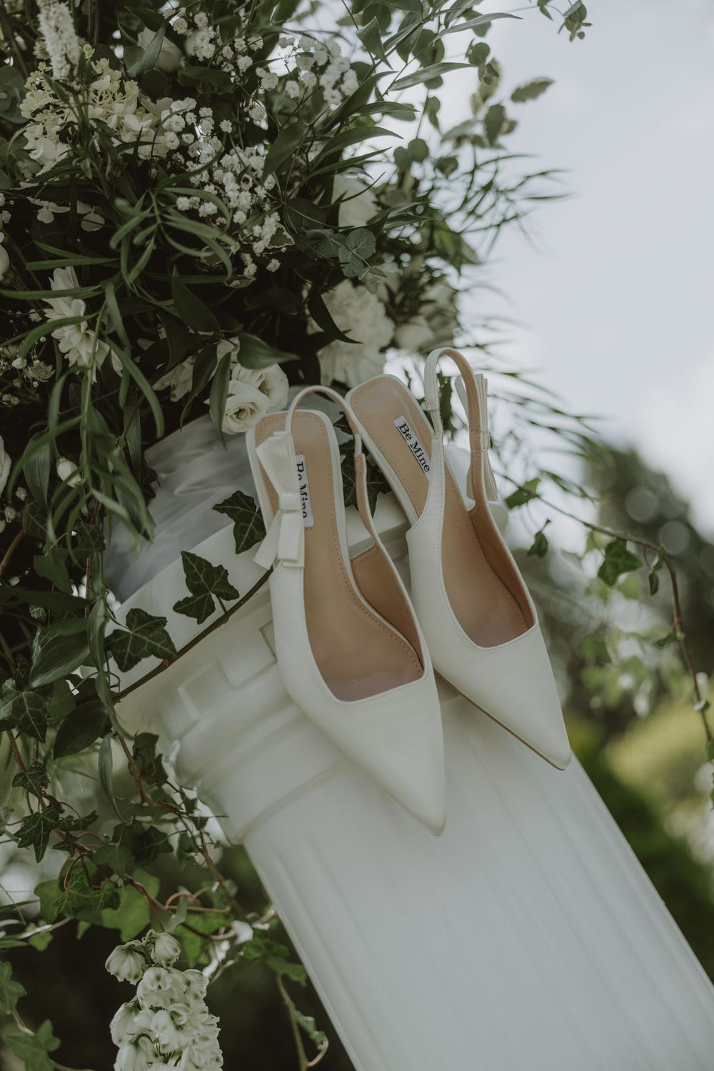Elegant white slingback bridal shoes displayed with floral arrangements and wedding dress fabric.