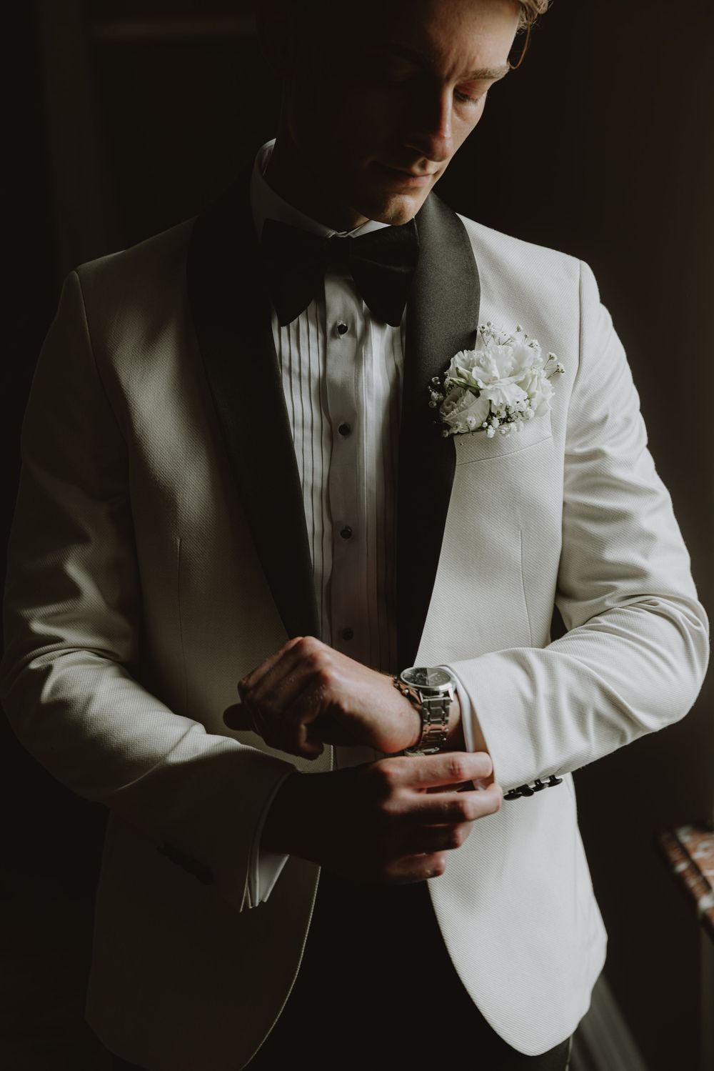 Close-up details of a groom in a white tuxedo jacket adjusting his cufflinks in dramatic lighting.