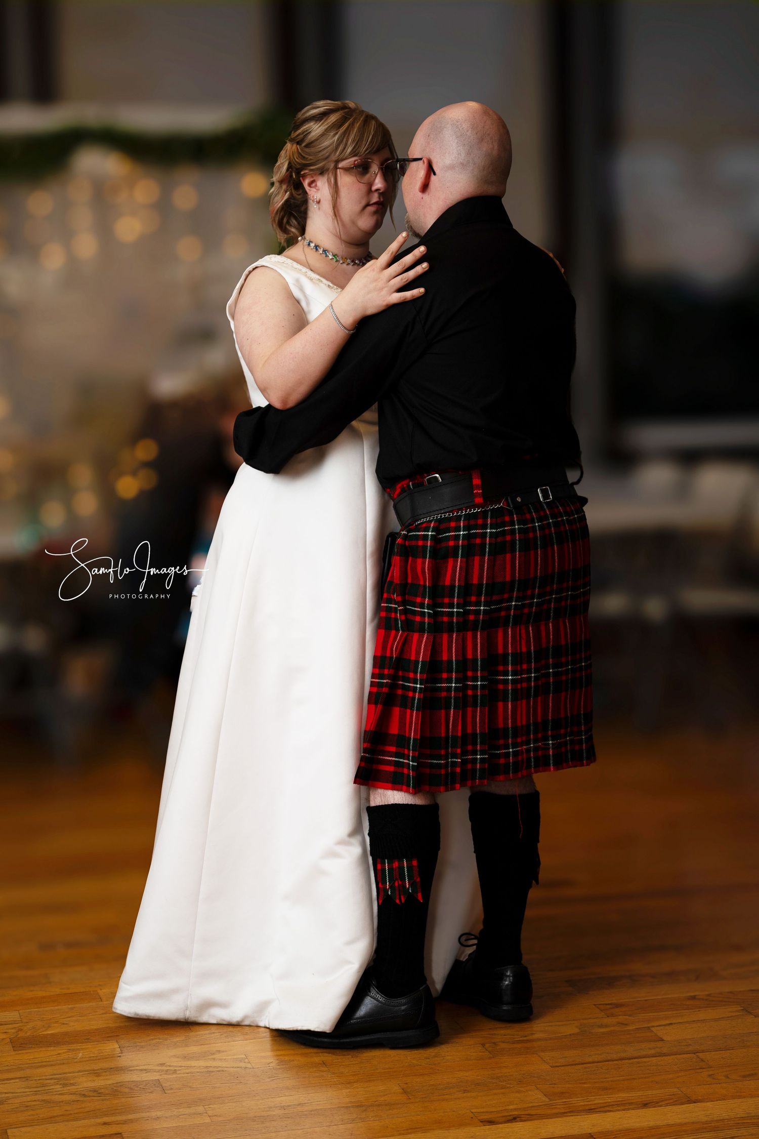 A couple shares a romantic wedding dance moment, with the groom wearing a traditional red and black tartan kilt.