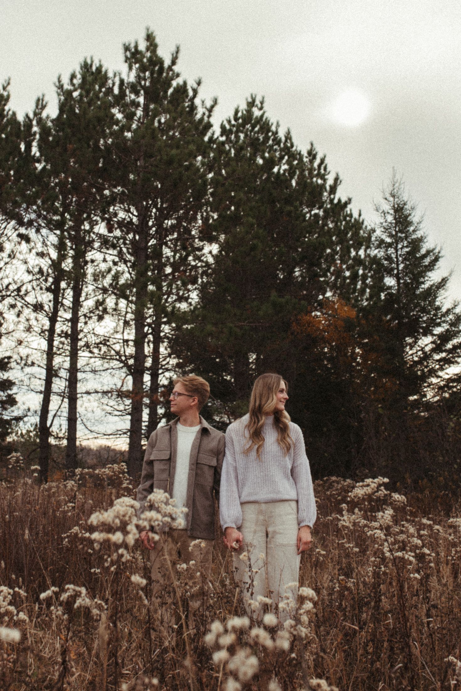 A couple standing in a field of autumn grass with tall evergreen trees and a cloudy sky behind them.
