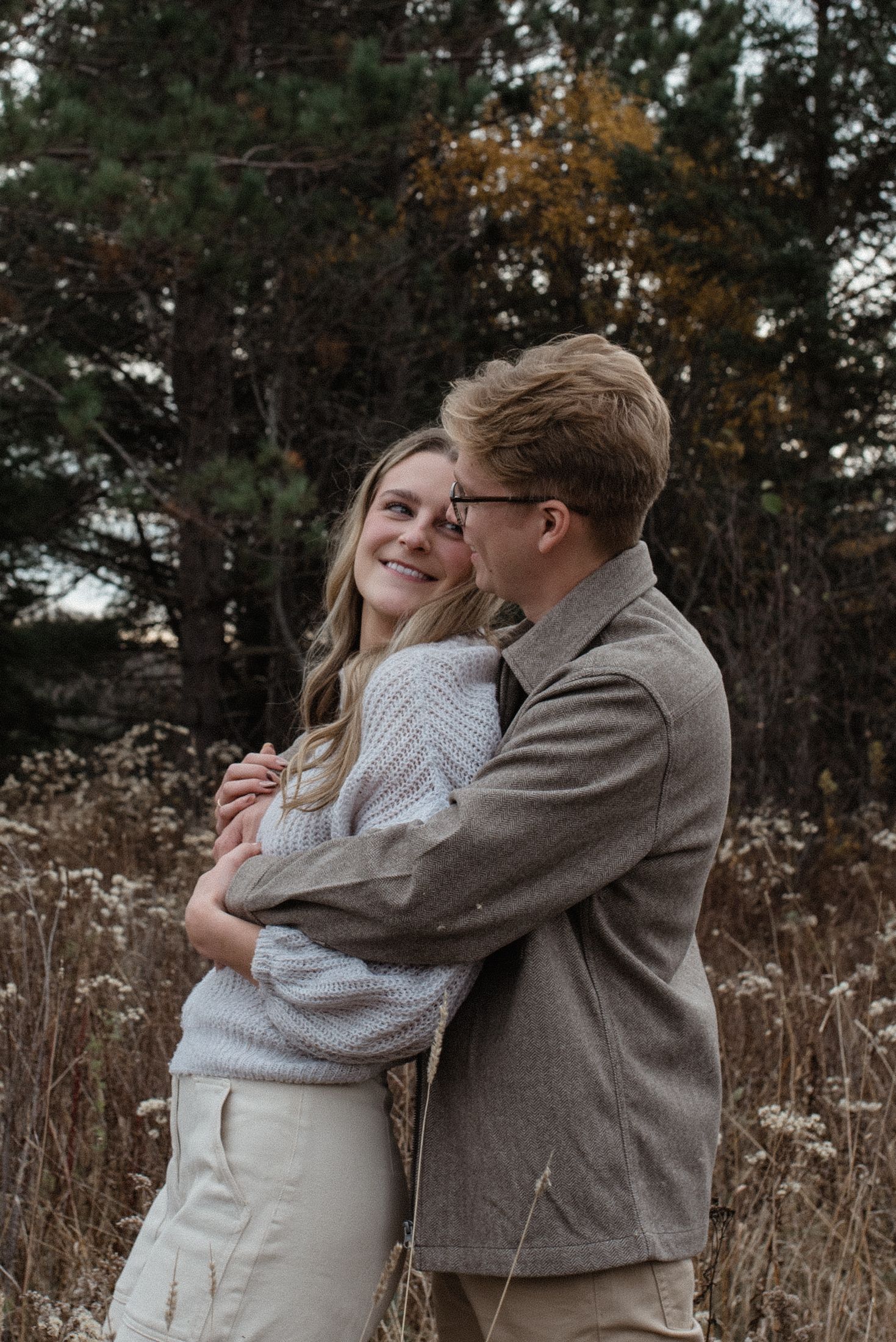 A couple embraces in a romantic autumn setting with tall grass and trees in the background during golden hour.
