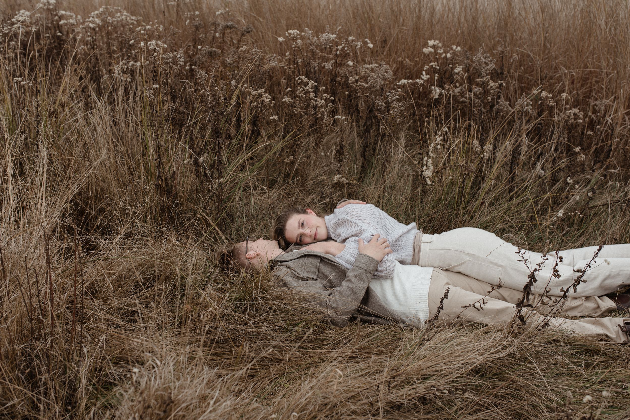A couple lying together in a field of dried tall grass and wildflowers on an overcast day.