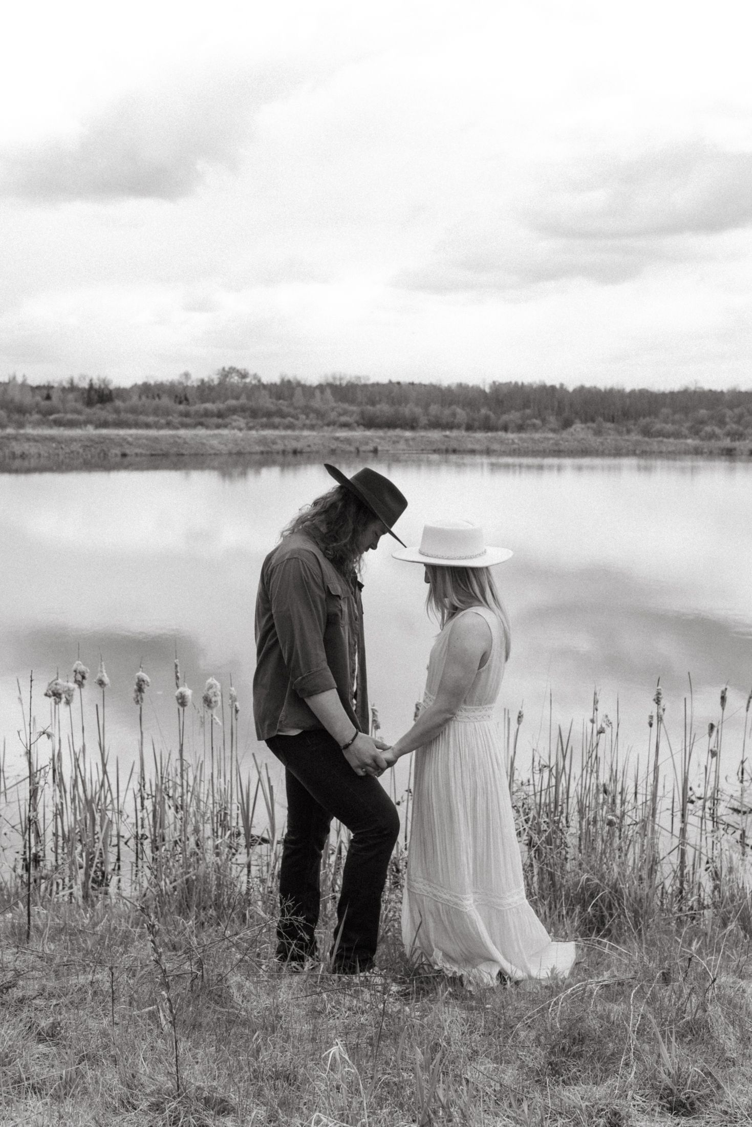A couple in western attire stands by a serene lake with cattails, captured in black and white photography.