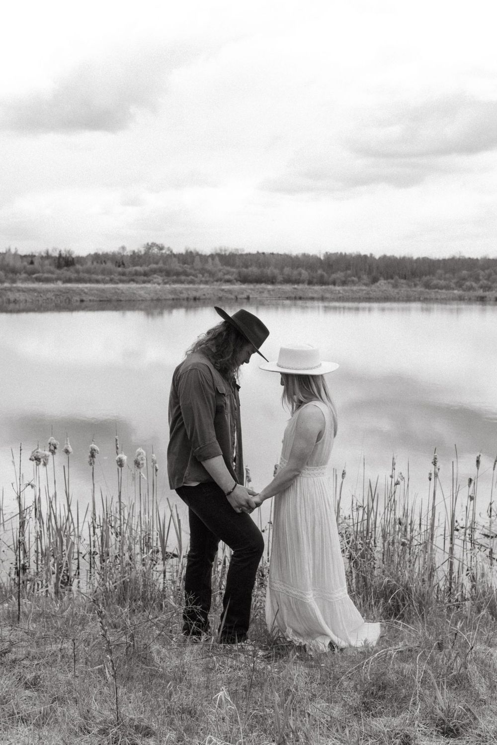 A couple in western attire stands by a serene lake with cattails, captured in black and white photography.