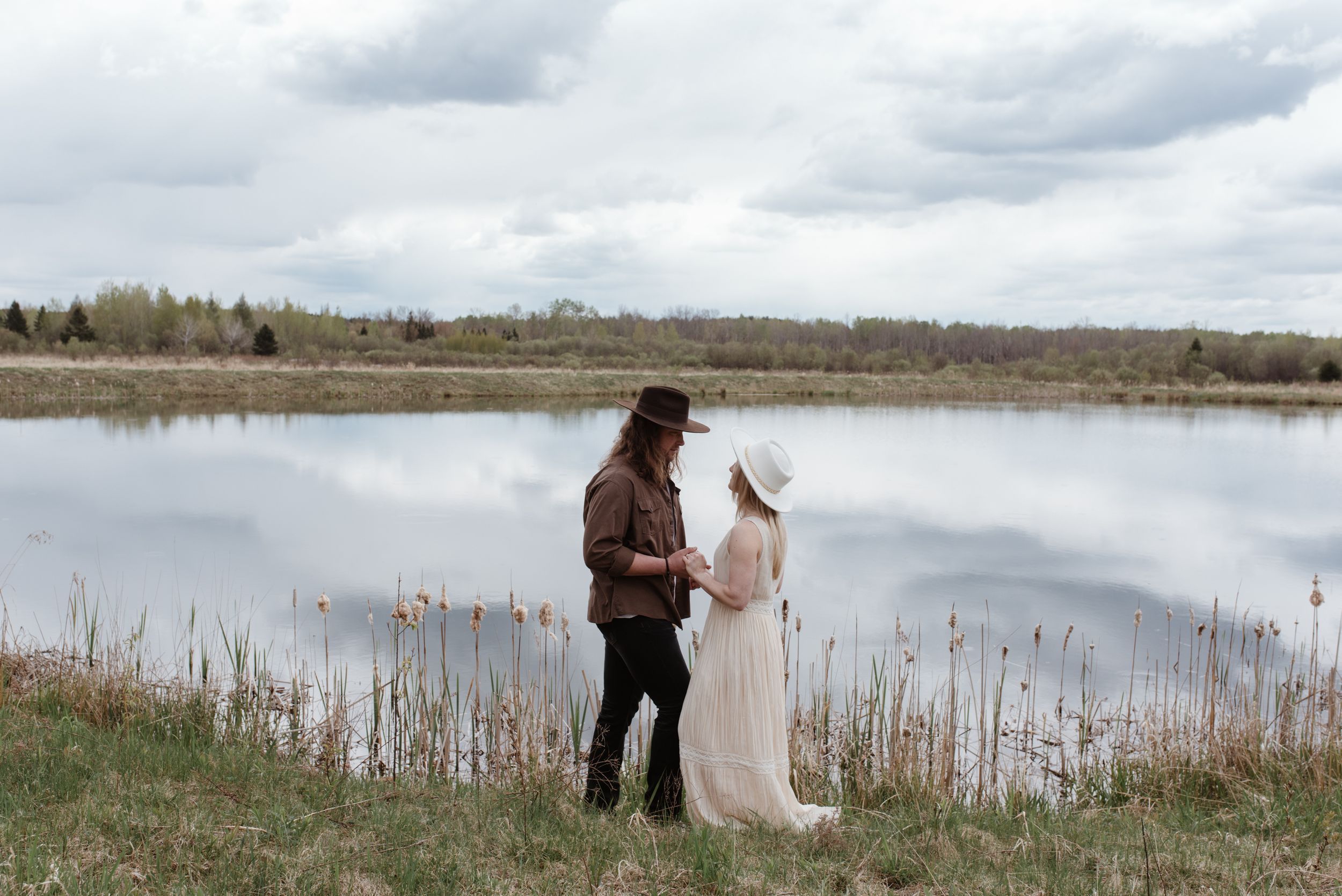 Two people standing by a serene lake with tall grass and trees under cloudy skies.