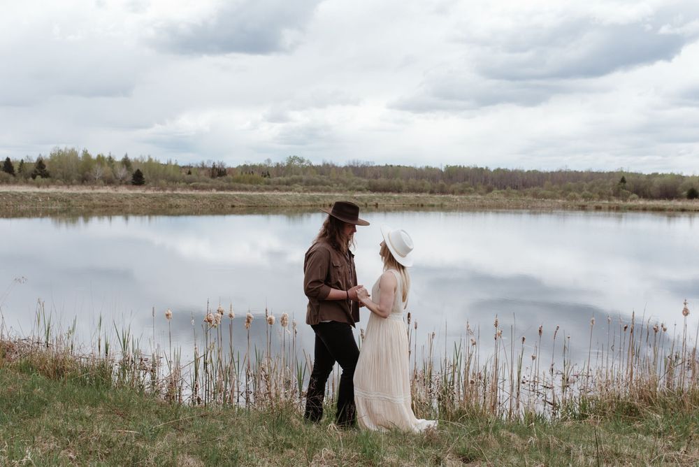 Two people standing by a serene lake with tall grass and trees under cloudy skies.