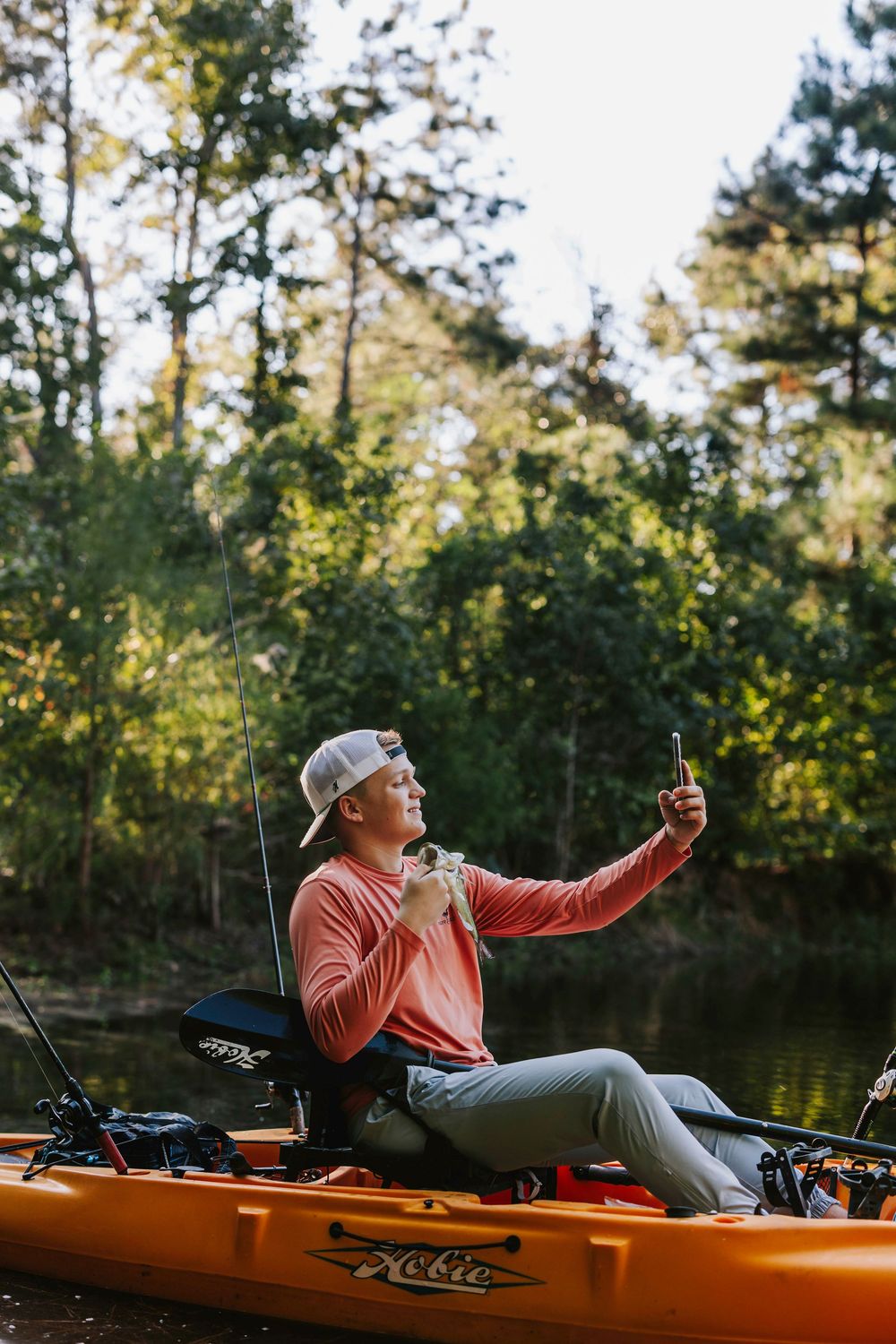 Person in orange shirt takes selfie while kayak fishing on peaceful water.