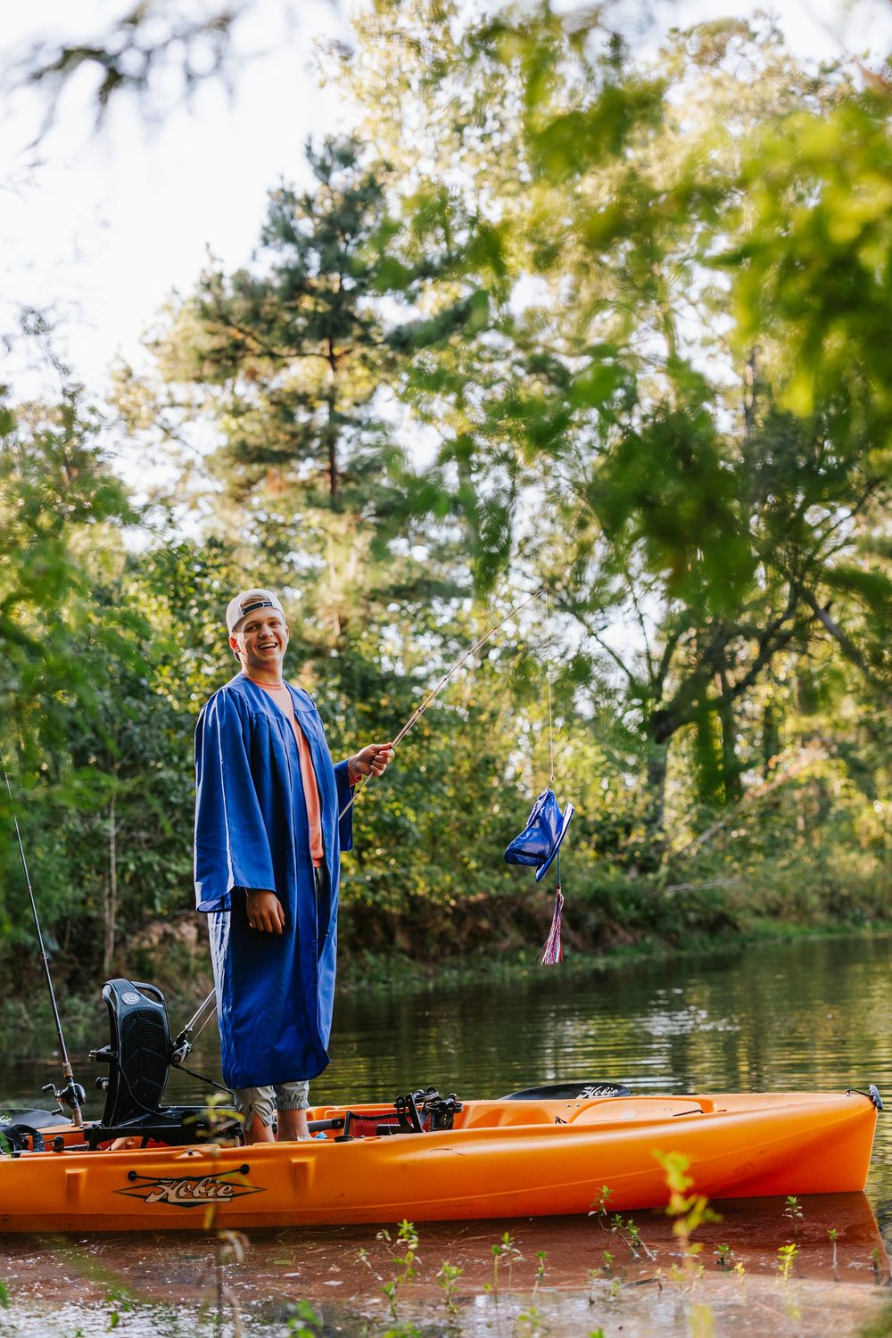 Person in blue graduation gown stands on orange kayak while fishing.