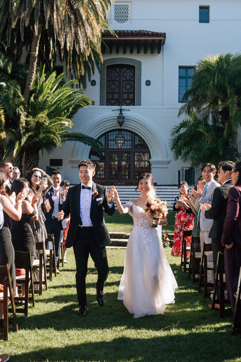 A bride and groom leave the aisle and smile after their wedding at the Sunken Gardens at the Santa Barbara Courthouse