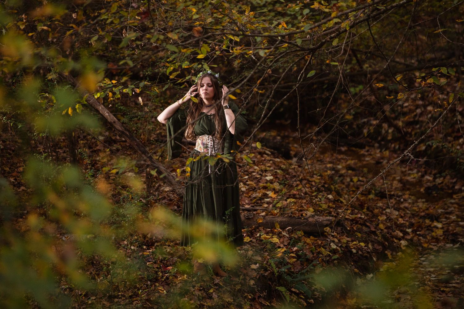 A person in vintage medieval dress stands in a forest clearing surrounded by autumn leaves.