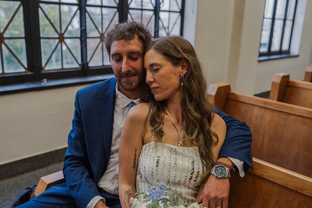 A couple shares an intimate moment while sitting in a church pew, the bride wearing a white dress and groom in navy suit.