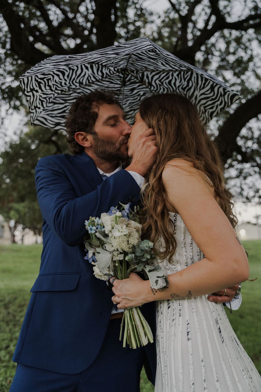 Newlyweds kiss under a zebra-striped umbrella while holding a white floral bouquet.