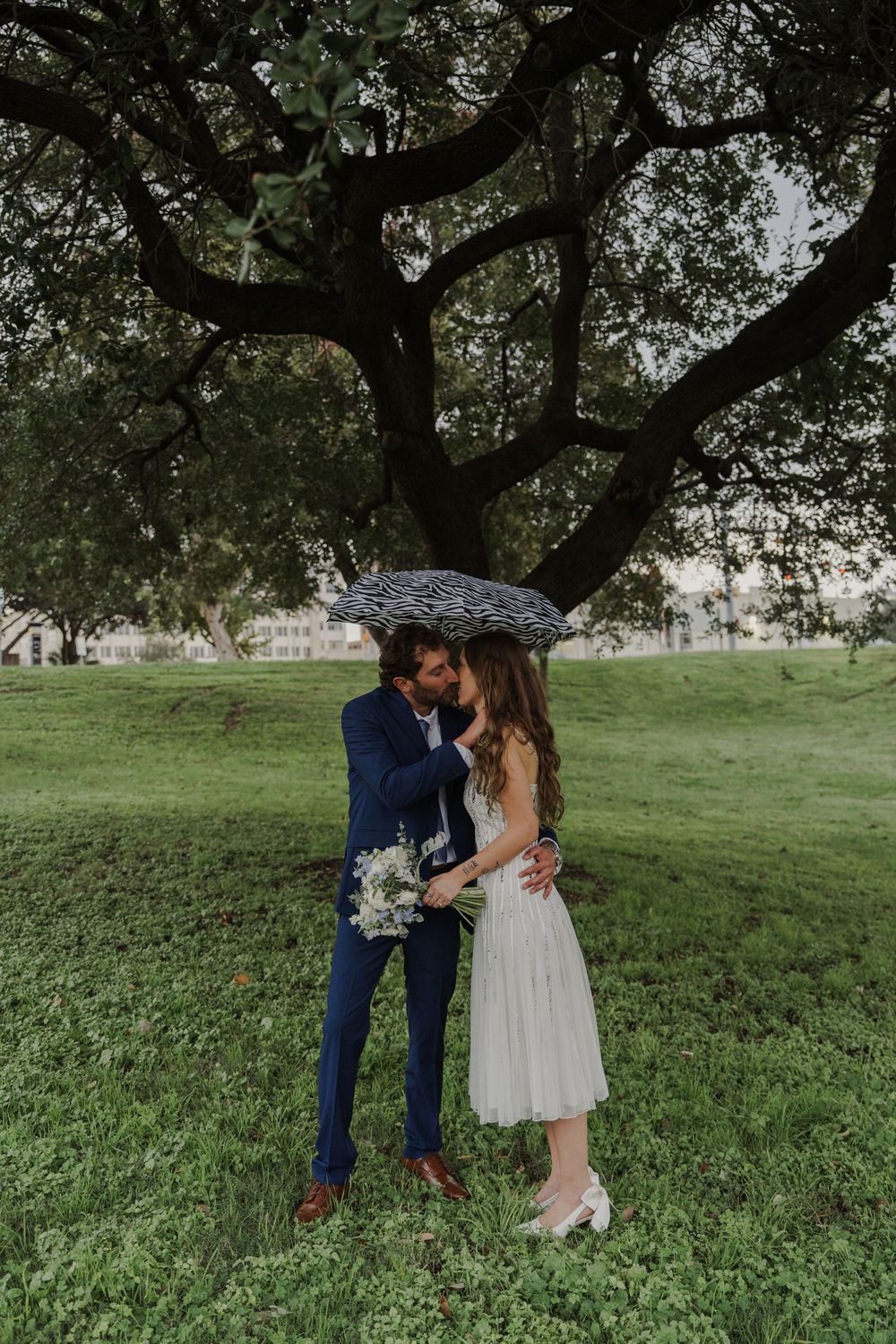 A couple in wedding attire stands beneath a large tree holding a zebra print umbrella.