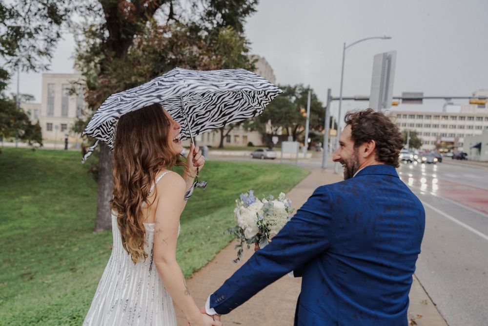 A couple in wedding attire walks through an urban park holding a zebra print umbrella.