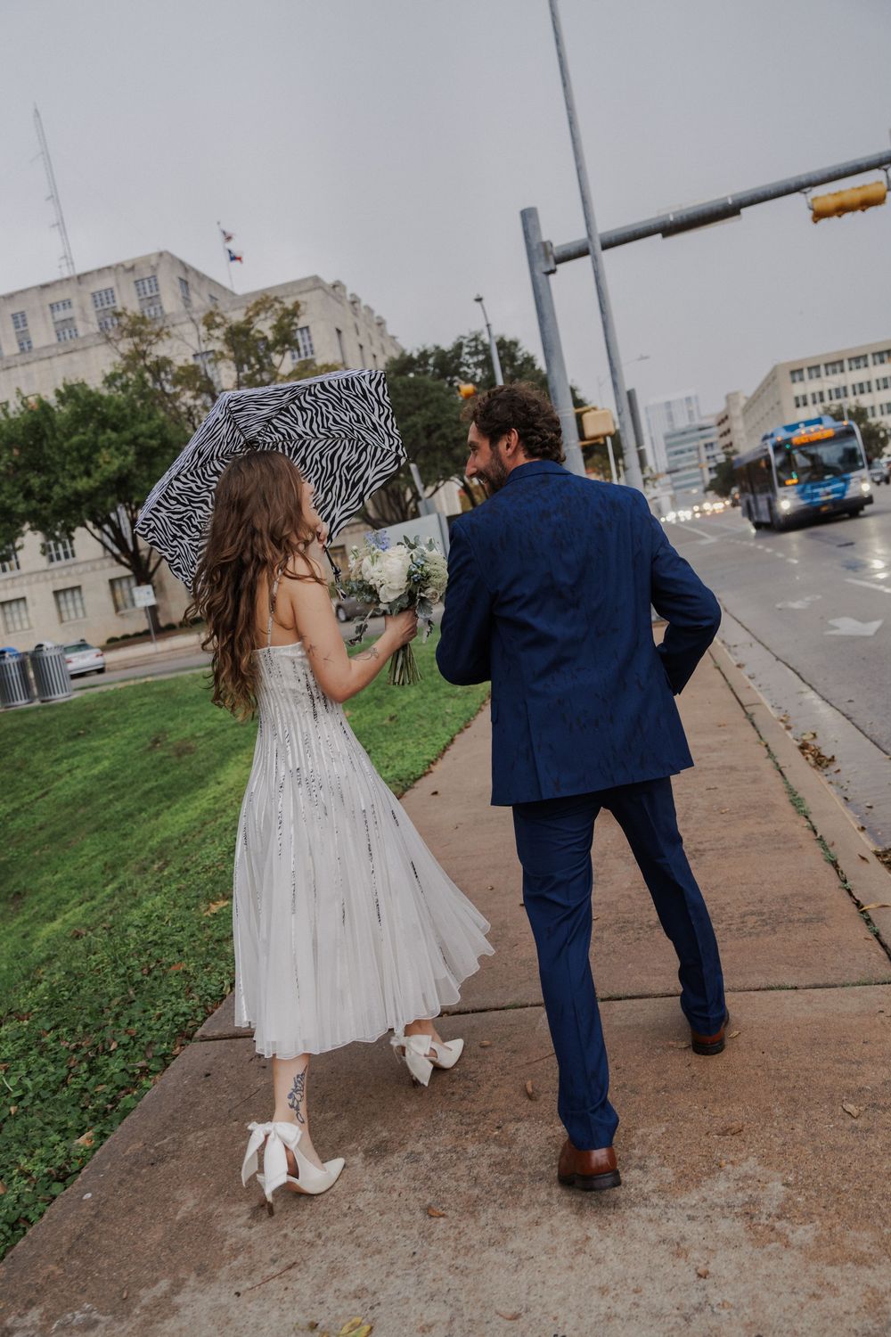 Bride and groom walk down a city sidewalk with a zebra print umbrella and white dress flowing.