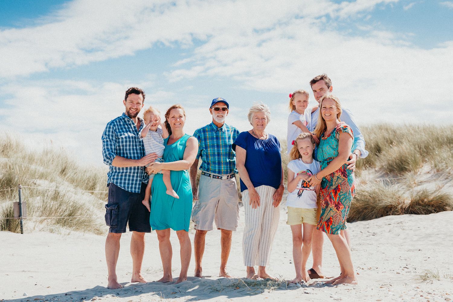 Multi-generational family group photo on sandy beach with dunes and blue sky.
