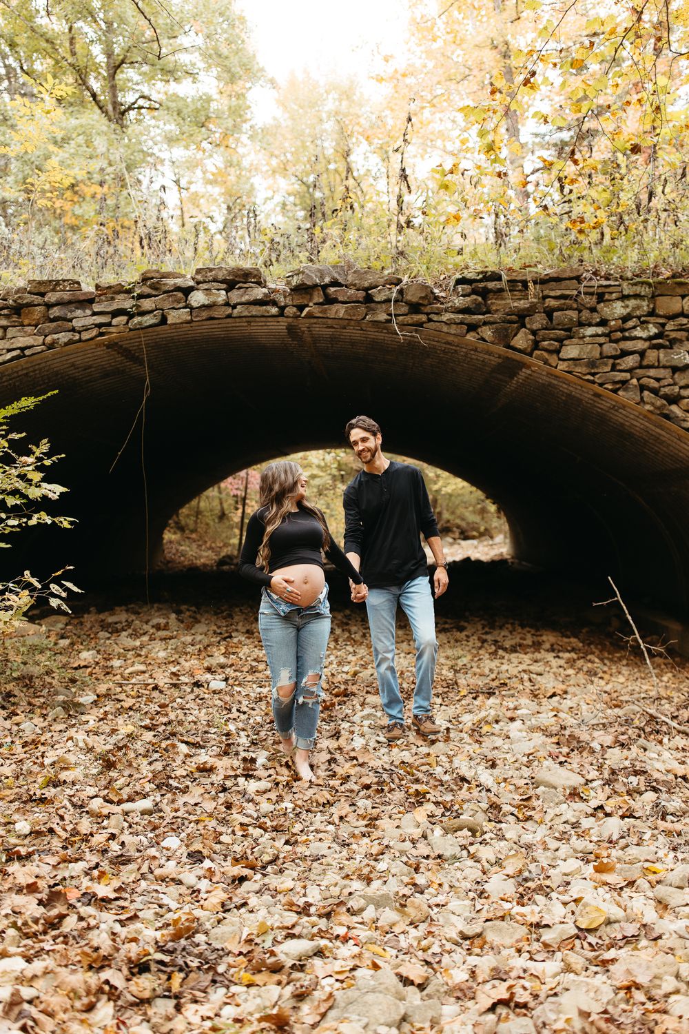 A couple walks together on fallen autumn leaves beneath a rustic stone bridge in a park setting.