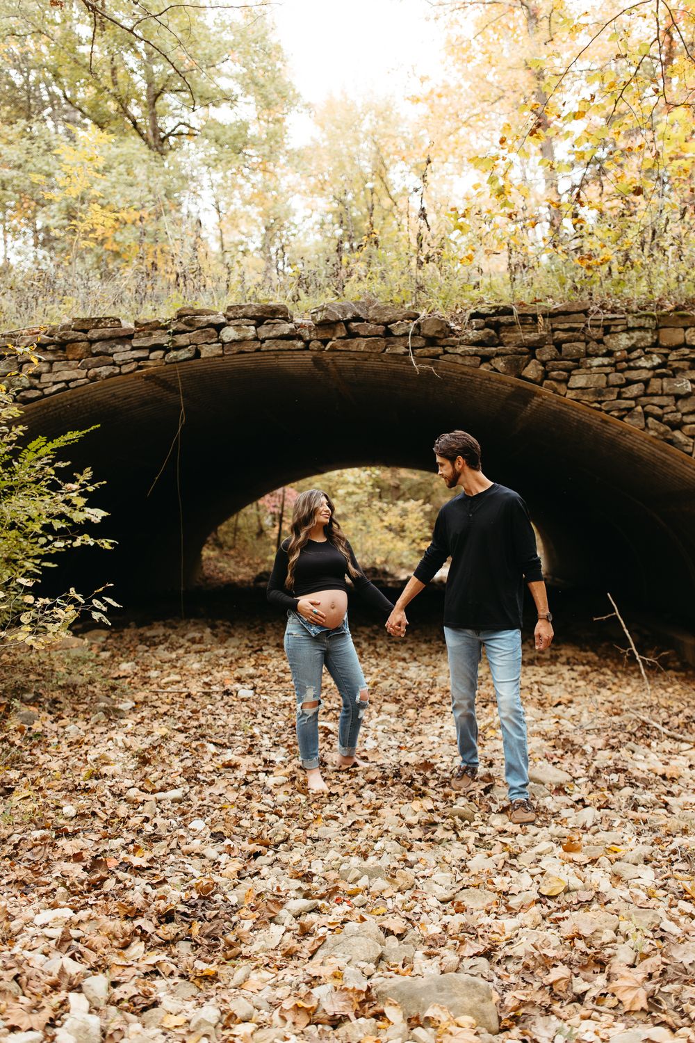 Two people in casual attire explore beneath a rustic stone bridge surrounded by fallen autumn leaves.