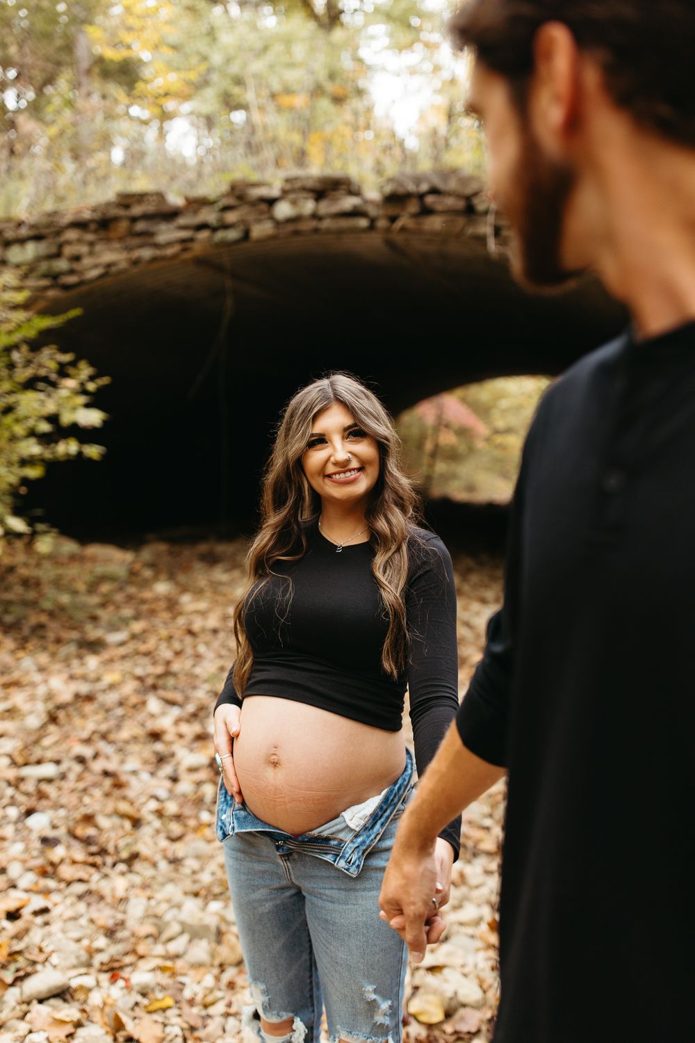 A couple shares a tender moment in autumn woods while showing off a pregnant belly in casual attire.