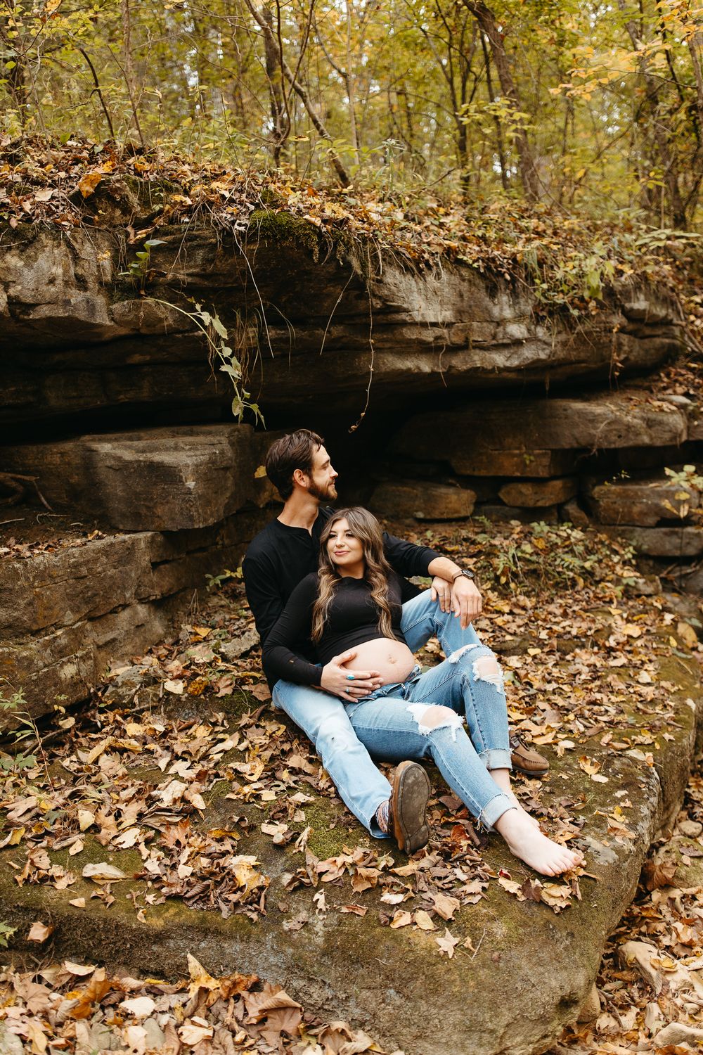 A couple relaxes together against a rocky outcrop surrounded by fallen autumn leaves in a natural outdoor setting.