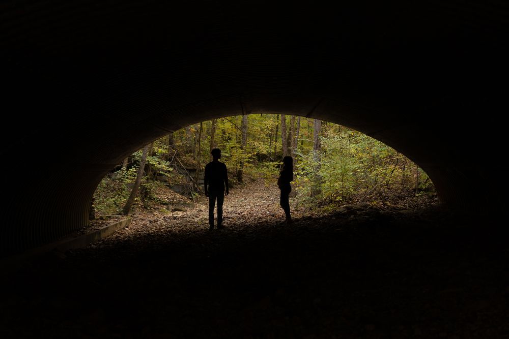 Two silhouetted figures walking through a dark tunnel with sunlit forest visible at the end.