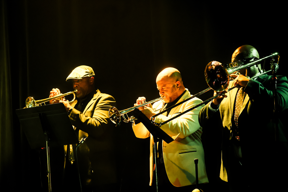 Musicians perform with brass instruments on a dark stage illuminated by dramatic spotlights during a jazz concert.