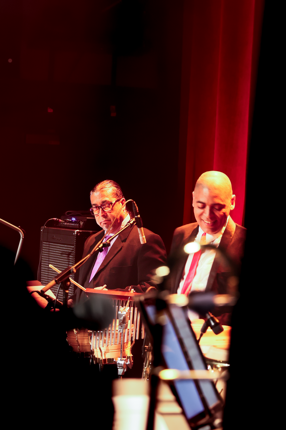 Musicians perform on stage in dramatic red lighting with instruments and equipment visible in the dark atmosphere.