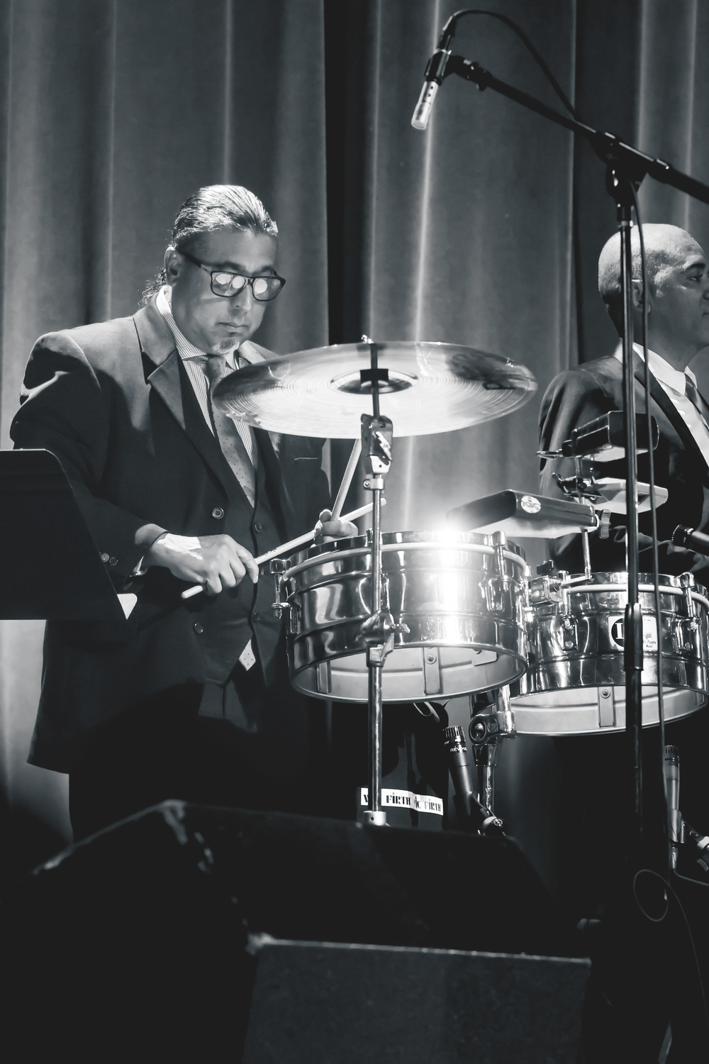 A drummer in a suit and glasses performs on stage with a drum kit and microphone setup in black and white.