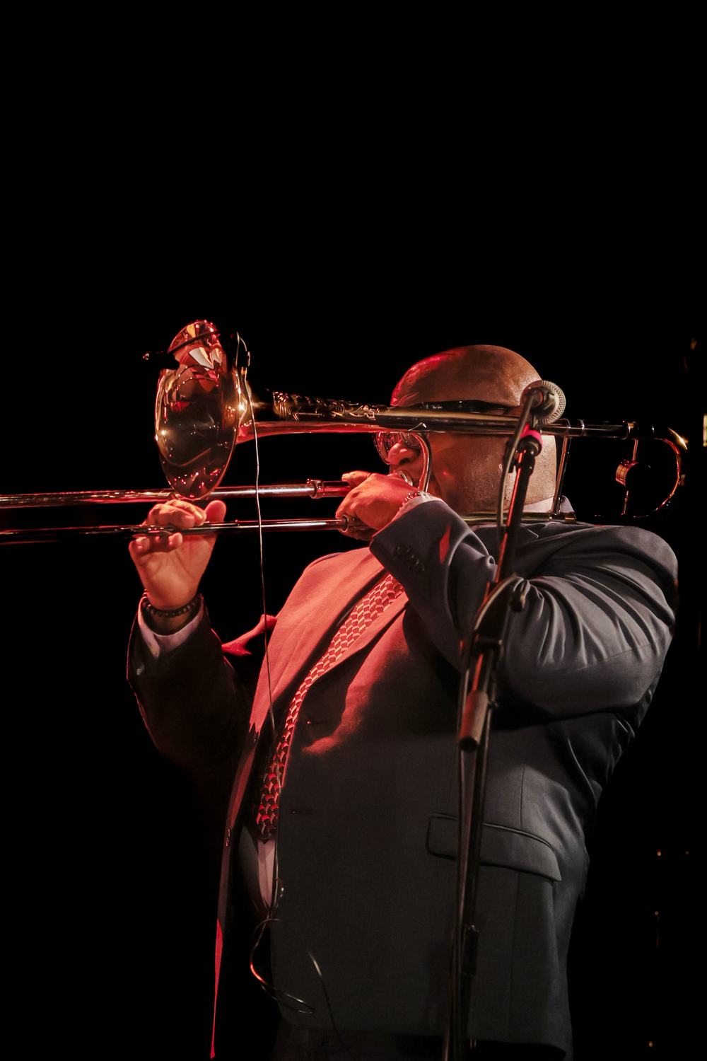 A musician in formal attire plays trombone on a dark stage illuminated by dramatic red lighting.