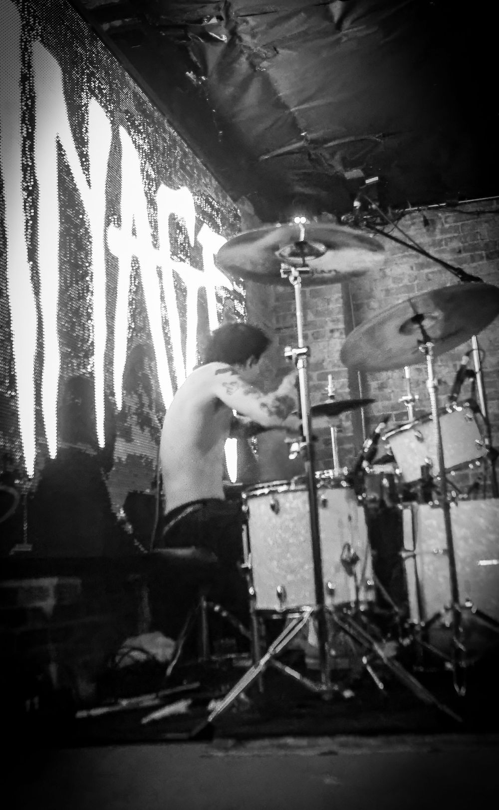 Shirtless drummer performs energetically on stage with dramatic lighting and band name illuminated behind the drum kit.