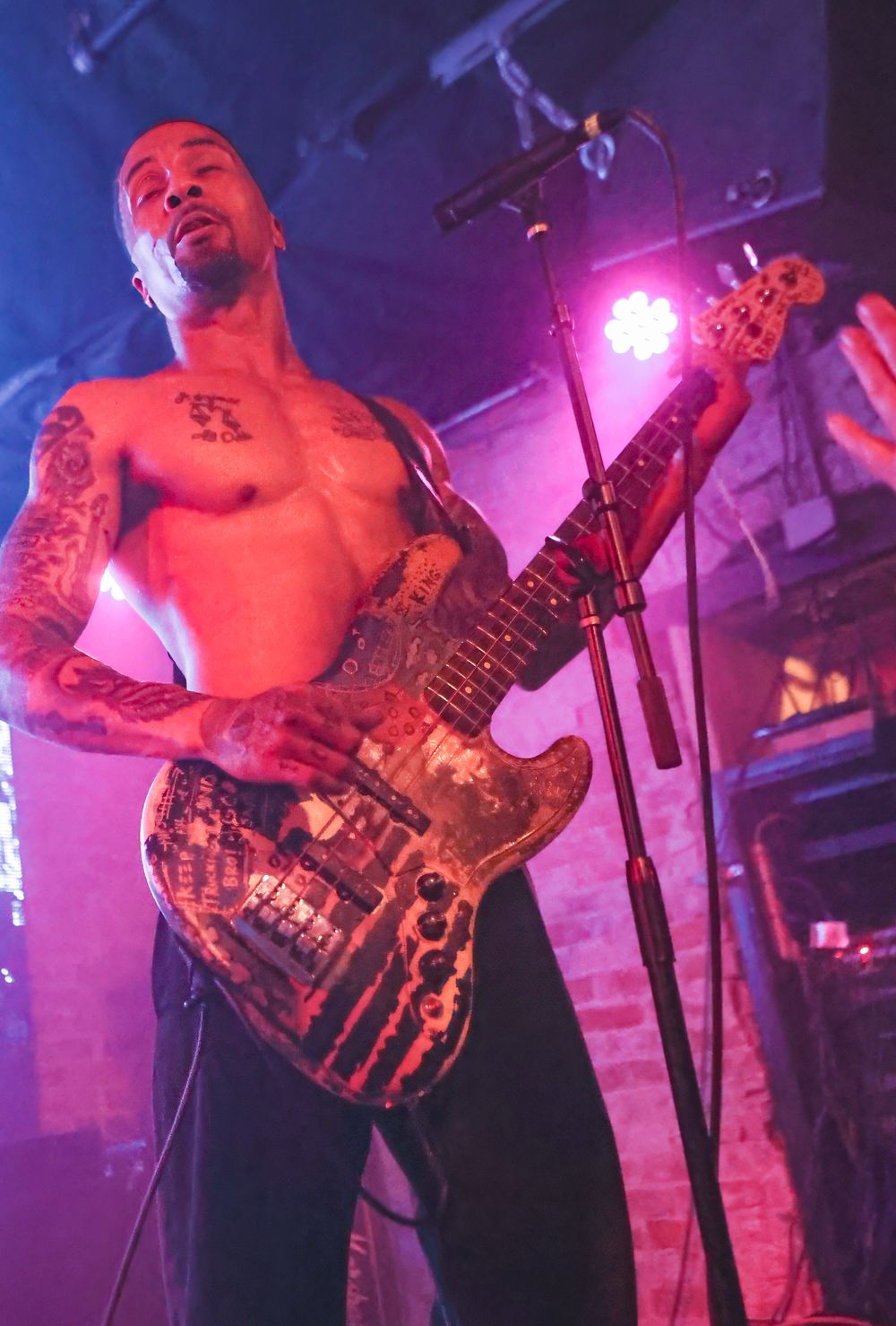 Shirtless musician performs on stage with guitar under dramatic purple and pink lighting at a rock concert venue.