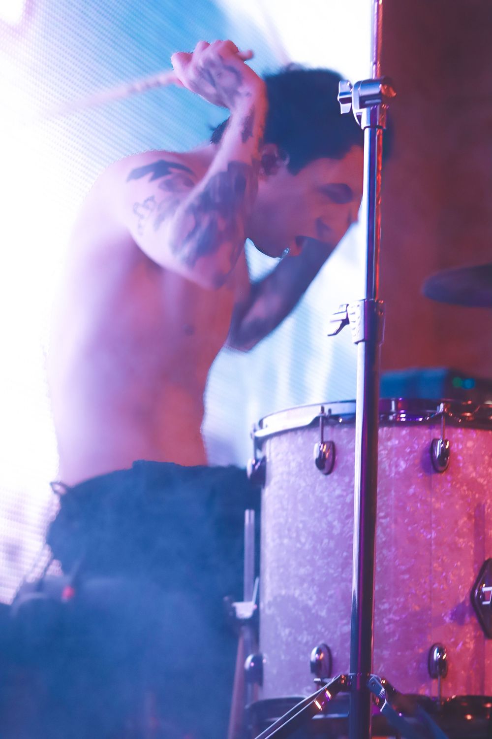 A drummer performs energetically on stage with sparkly drum kit in pink and blue lighting atmosphere.